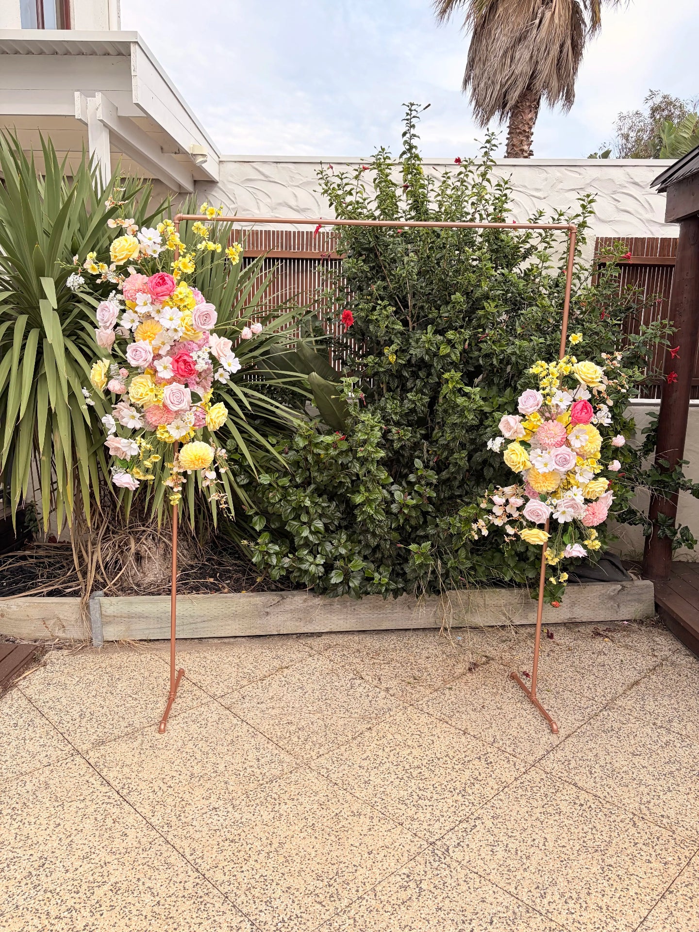 Two floral arrangements on stands in a garden setting with a palm tree in the background.