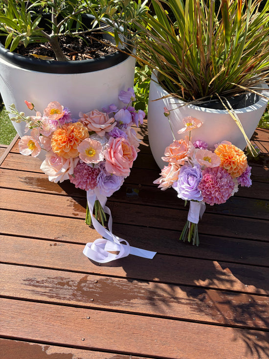 Two artificial wedding bouquets on a wooden surface with potted plants in the background.