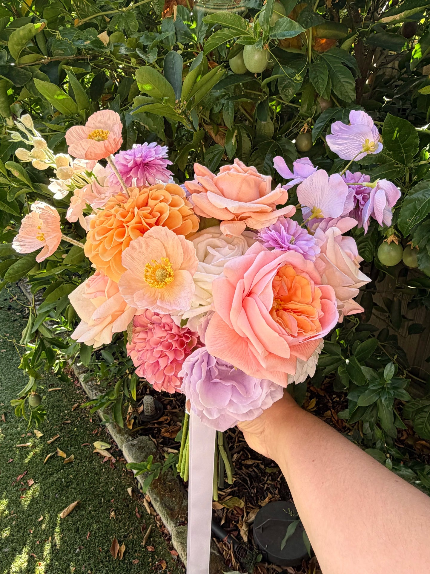 Faux wedding bouquet of pastel flowers held by a hand in front of green foliage