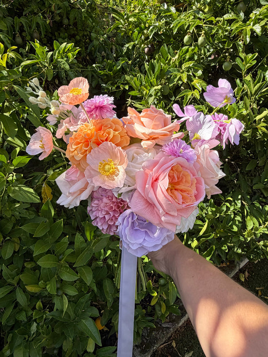 Hand holding a peach and lilac spring wedding bouquet with a green bush in the background