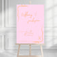 Pink party welcome sign with orange text on an easel against a white wall.
