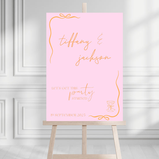Pink party welcome sign with orange text on an easel against a white wall.