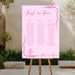 Pink seating chart on a wooden stand outdoors with plants and stone wall in the background