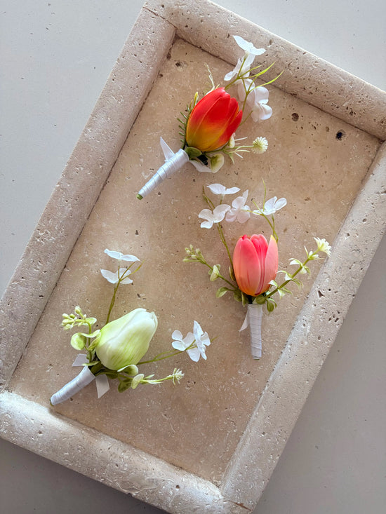 3 tulip buttonholes in orange, pink and white, with whimsical white wildflowers displayed on a travertine tray.