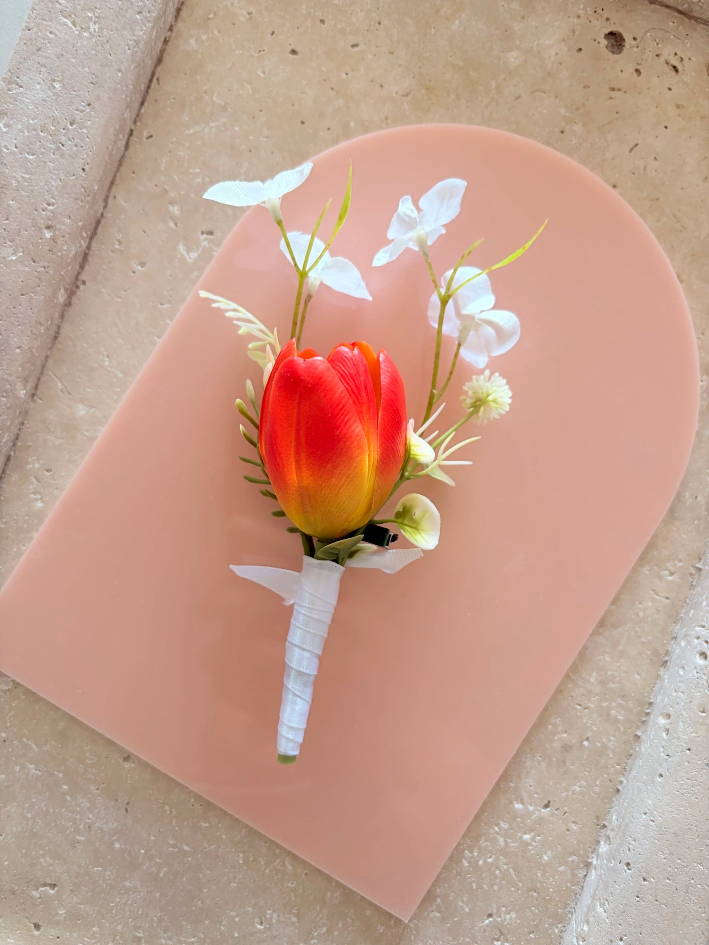 Orange buttonhole with whimsical white wildflowers displayed on a peach acrylic arch for product photography
