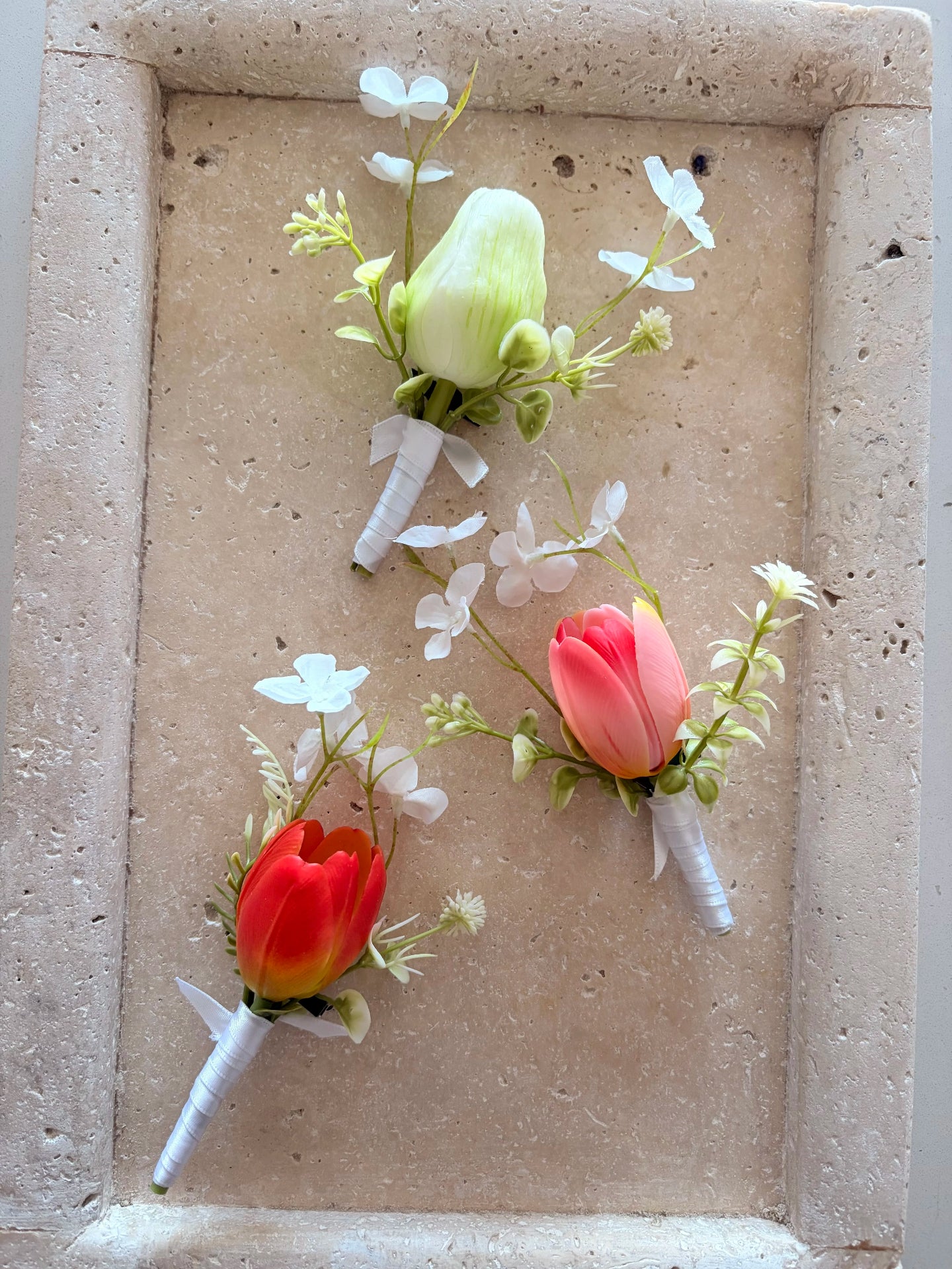 Group of three buttonholes in white, pink and orange with whimsical white wildflowers displayed on a travertine tray for product photography