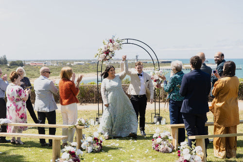 Ocean front wedding ceremony with a bride and groom under a floral arch, guests on either side.