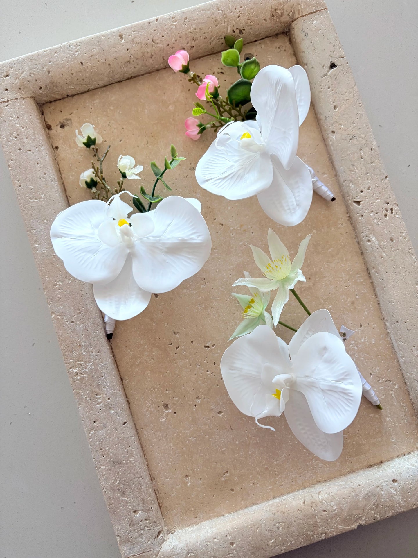 3 white orchid buttonholes, each with different detailing, on a textured tray.