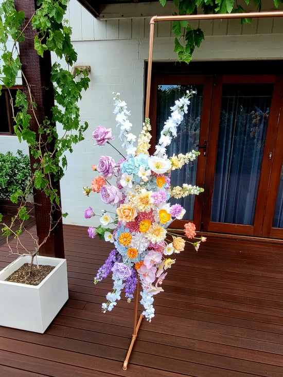 Colourful floral arrangement on a stand outdoors with a wooden deck and white wall background