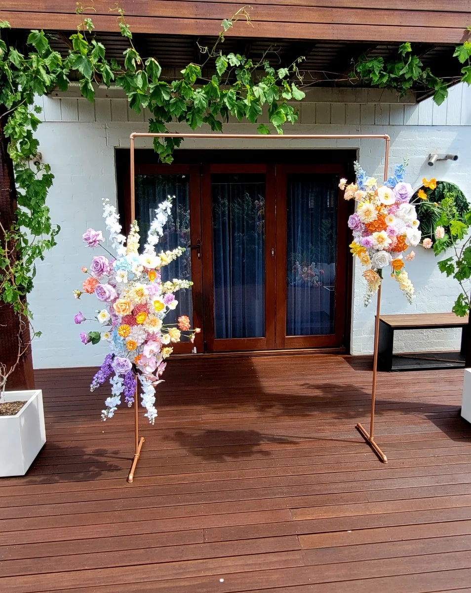 Two floral arrangements on stands in front of a building with glass doors with wooden deck and greenery.