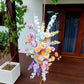 Colorful floral arrangement on a stand outdoors with a wooden deck and white wall background.