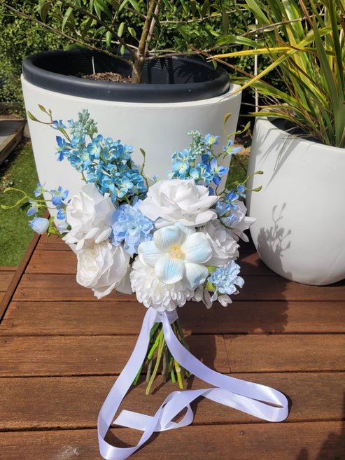 Bouquet of white and blue flowers with a ribbon on a wooden surface, with potted plants in the background.