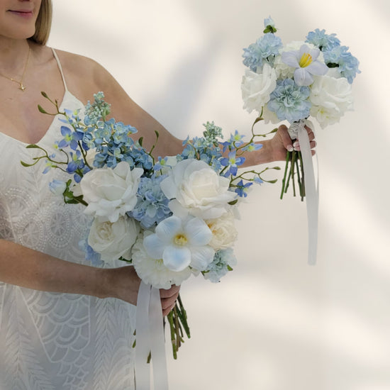 Woman holding two wedding bouquets of white and blue flowers against a plain background