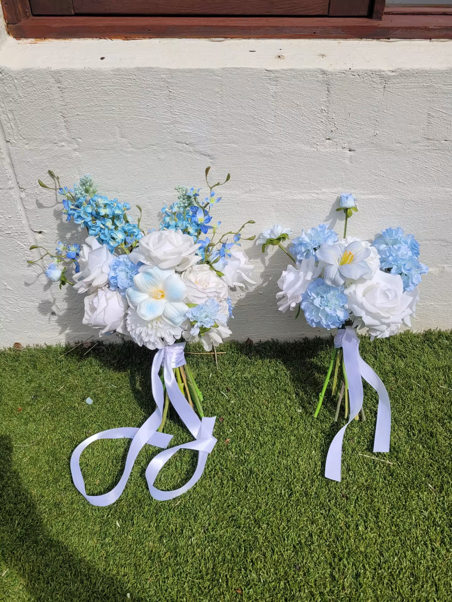 Two floral bouquets with ribbons on a grassy surface against a concrete wall.