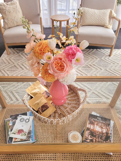 Floral arrangement in a pink vase on a glass coffee table with magazines and a basket.