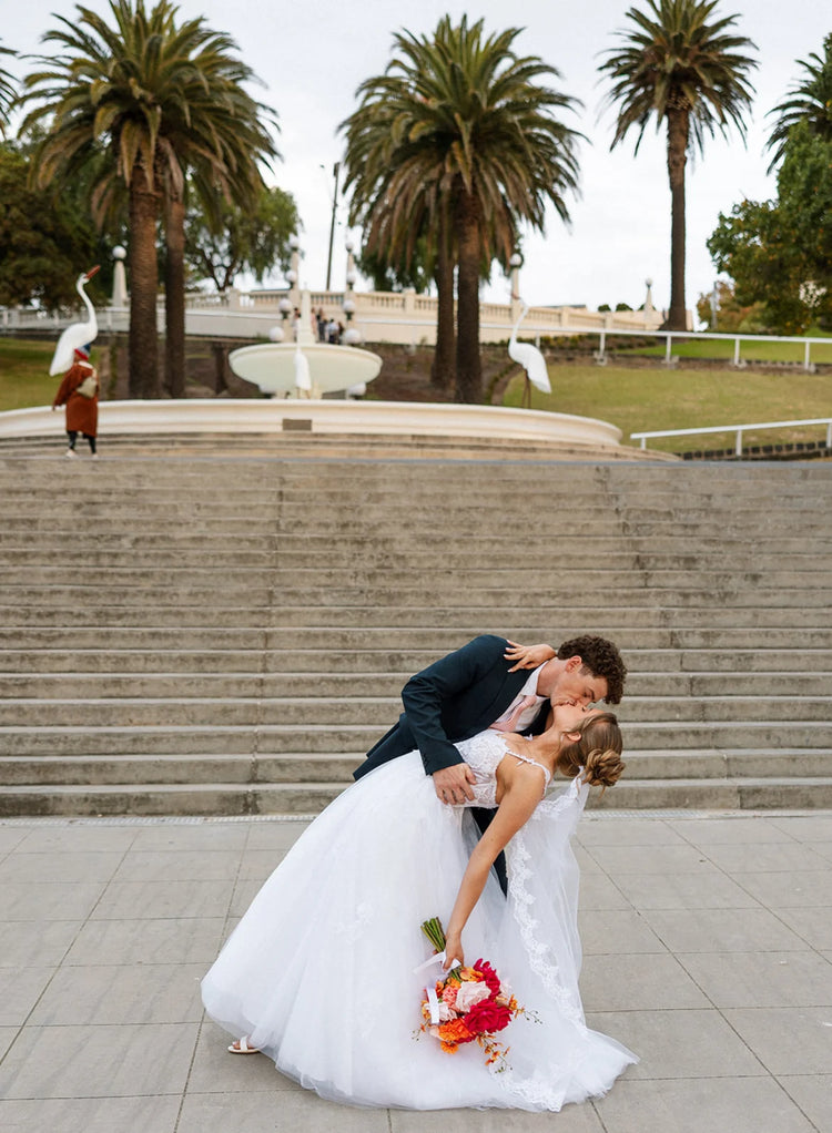 Couple in wedding attire and wedding bouquet doing 'the dip' on steps with palm trees and a fountain in the background
