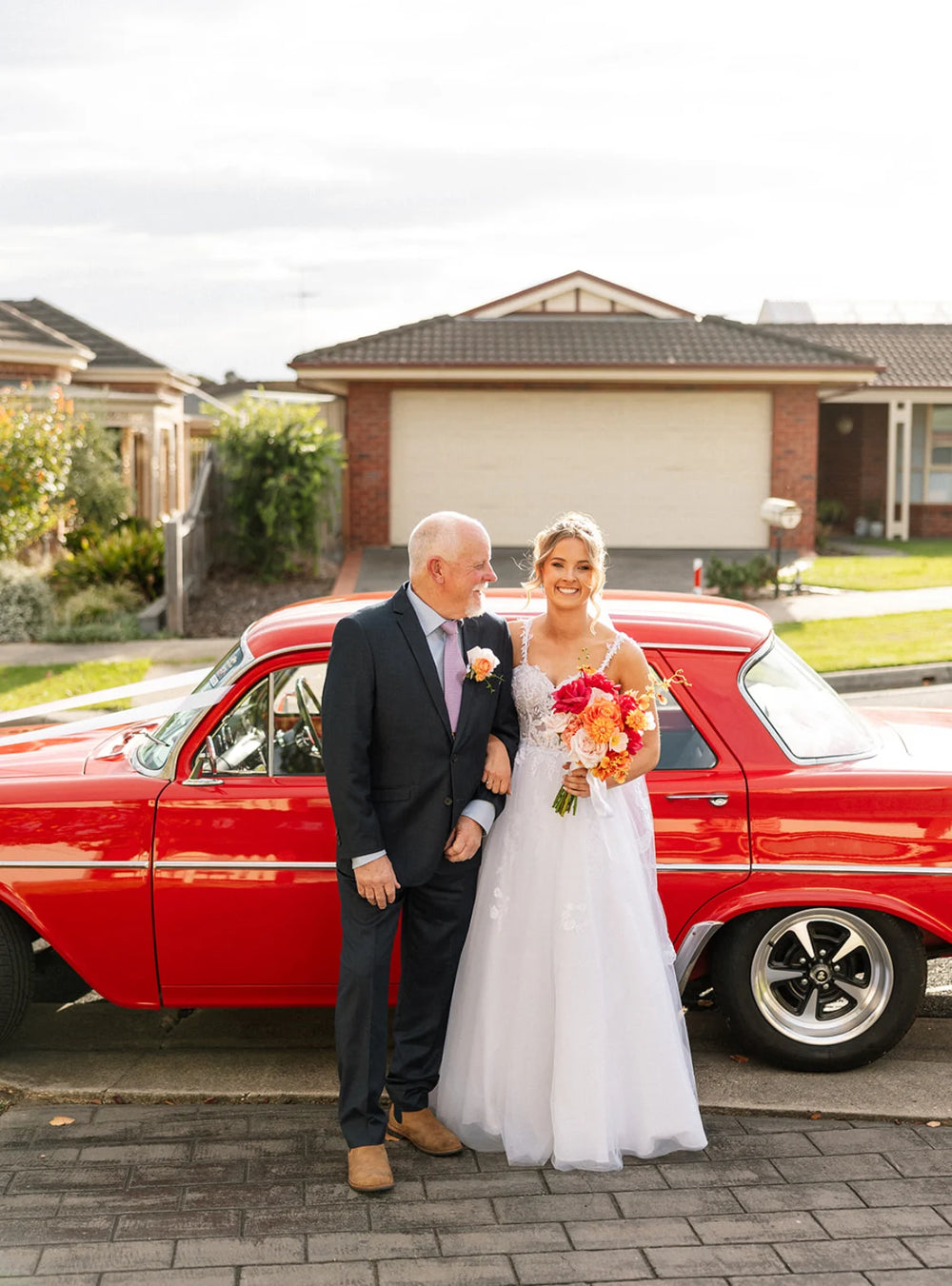 Bride with her wedding bouquet and father standing in front of a red vintage car on a residential street