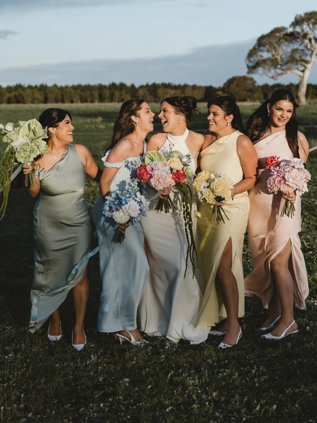 Bride and bridesmaids in formal dresses with pastel bouquets standing in a field.