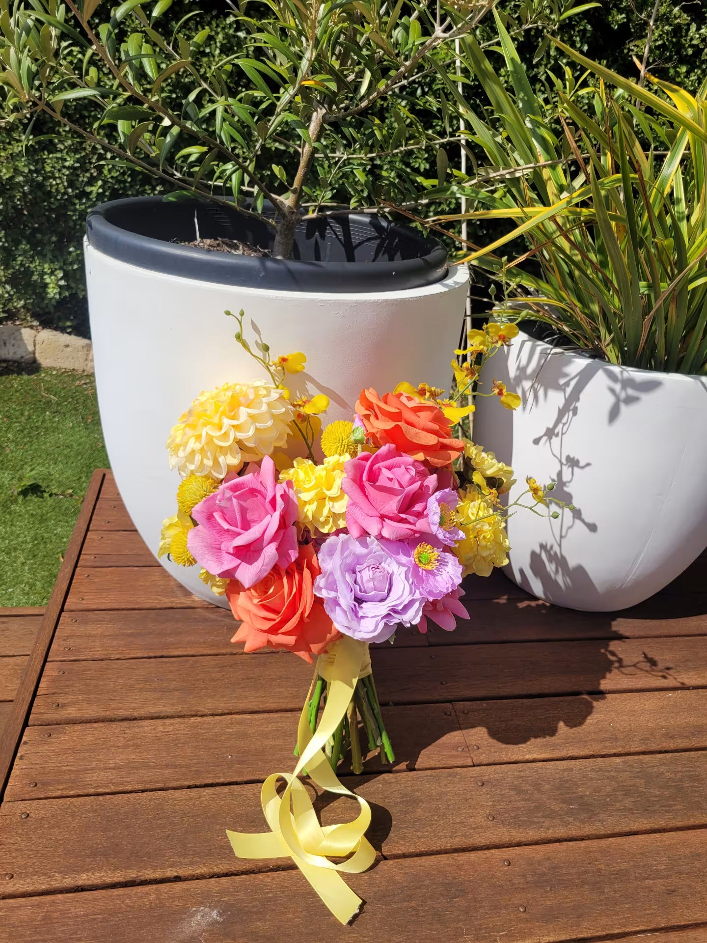 Wedding bouquet of colorful flowers in a white pot on a wooden deck with plants in the background.