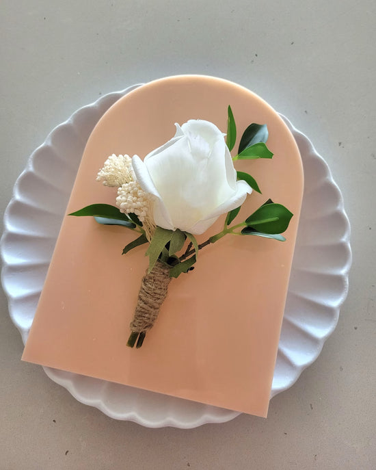 White rose boutonniere on a pink card with green leaves, placed on a white plate.