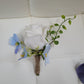Floral boutonniere with white rose, blue flowers, and green leaves on a white surface.