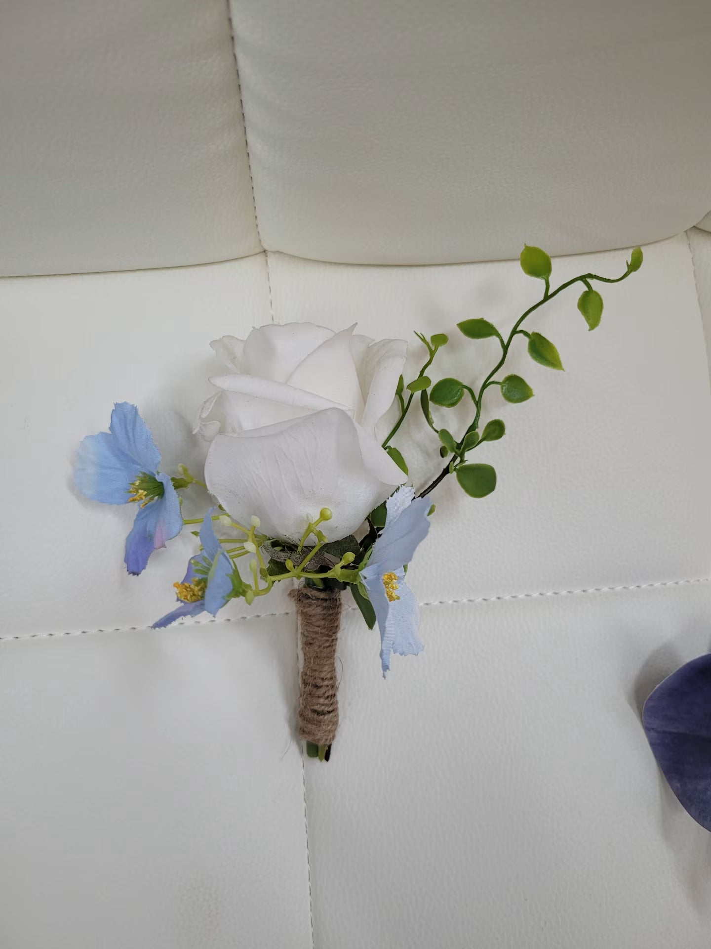 Floral boutonniere with white rose, blue flowers, and green leaves on a white surface.