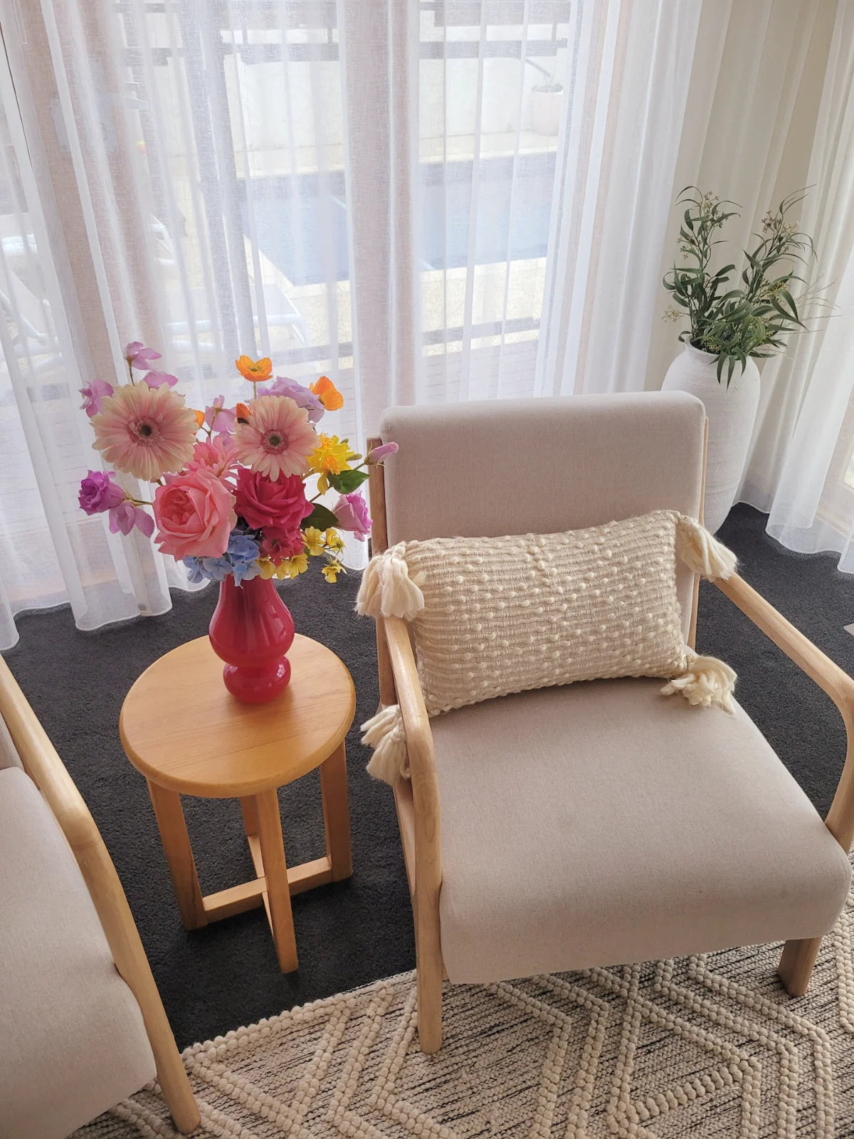 Beige armchair with a textured pillow, small round table with a pink vase and flowers, and white curtains in a room.