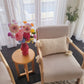 Beige armchair with a textured pillow, small round table with a pink vase and flowers, and sheer curtains in a bright room.