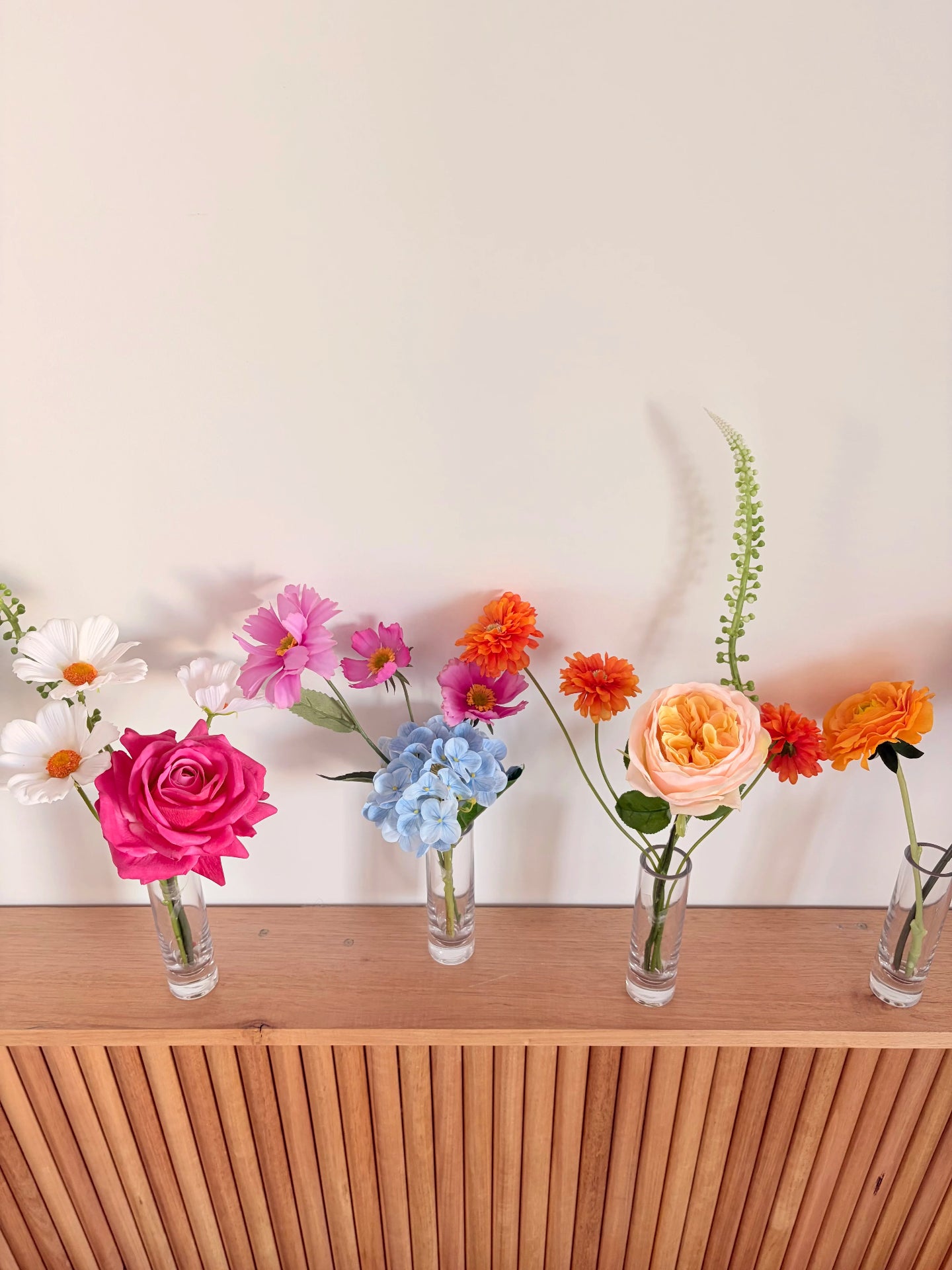 Five bud vases with colourful flower arrangements on a wooden surface against a white wall