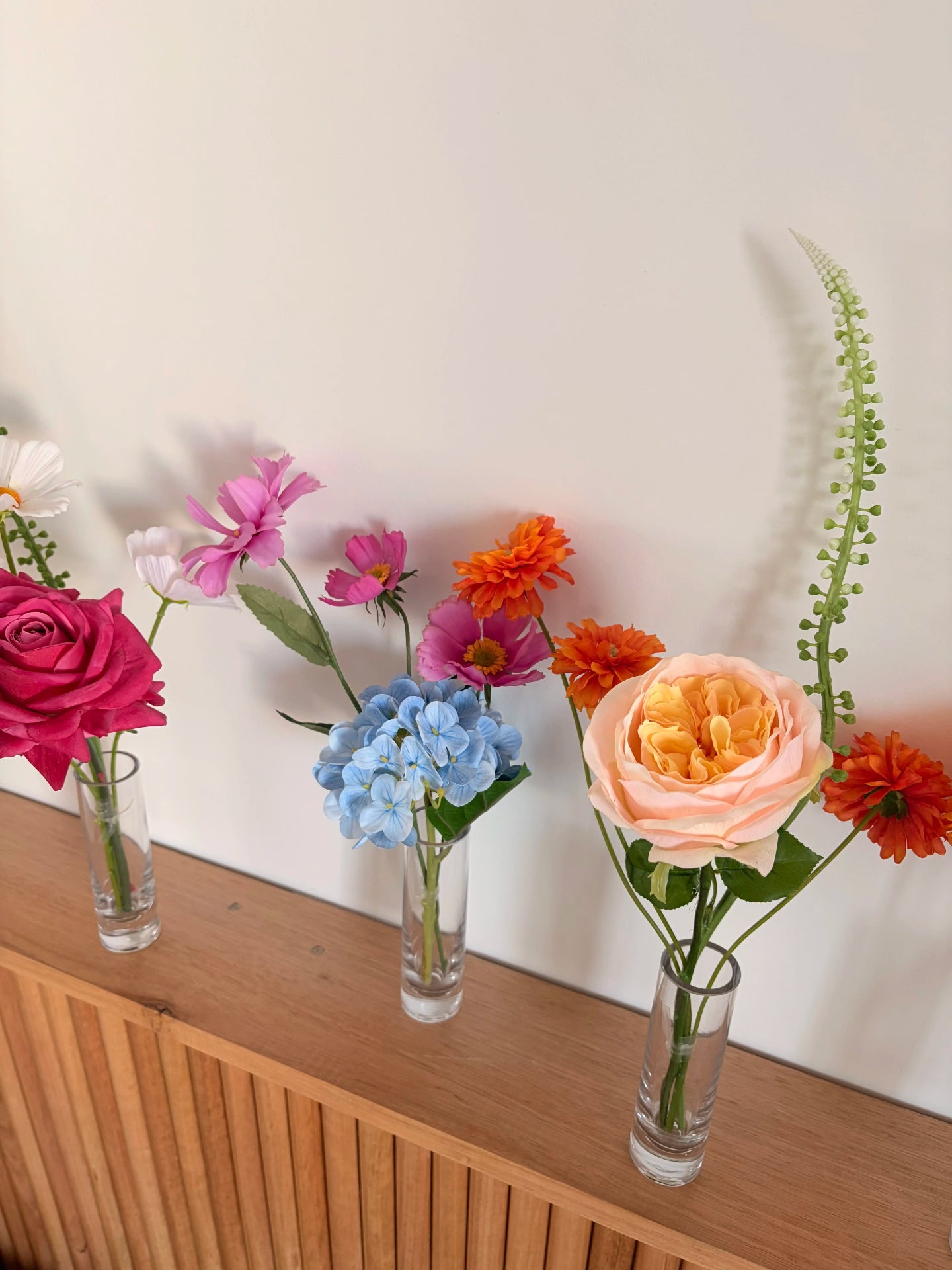 Colourful  faux flowers in clear vases on a wooden surface with a white wall background