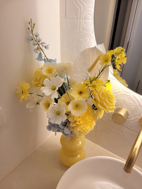 Yellow and white floral arrangement in a yellow vase on a bathroom counter.