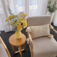 Beige armchair with a textured pillow, small round table with a yellow vase and flowers, and sheer curtains in a bright room.