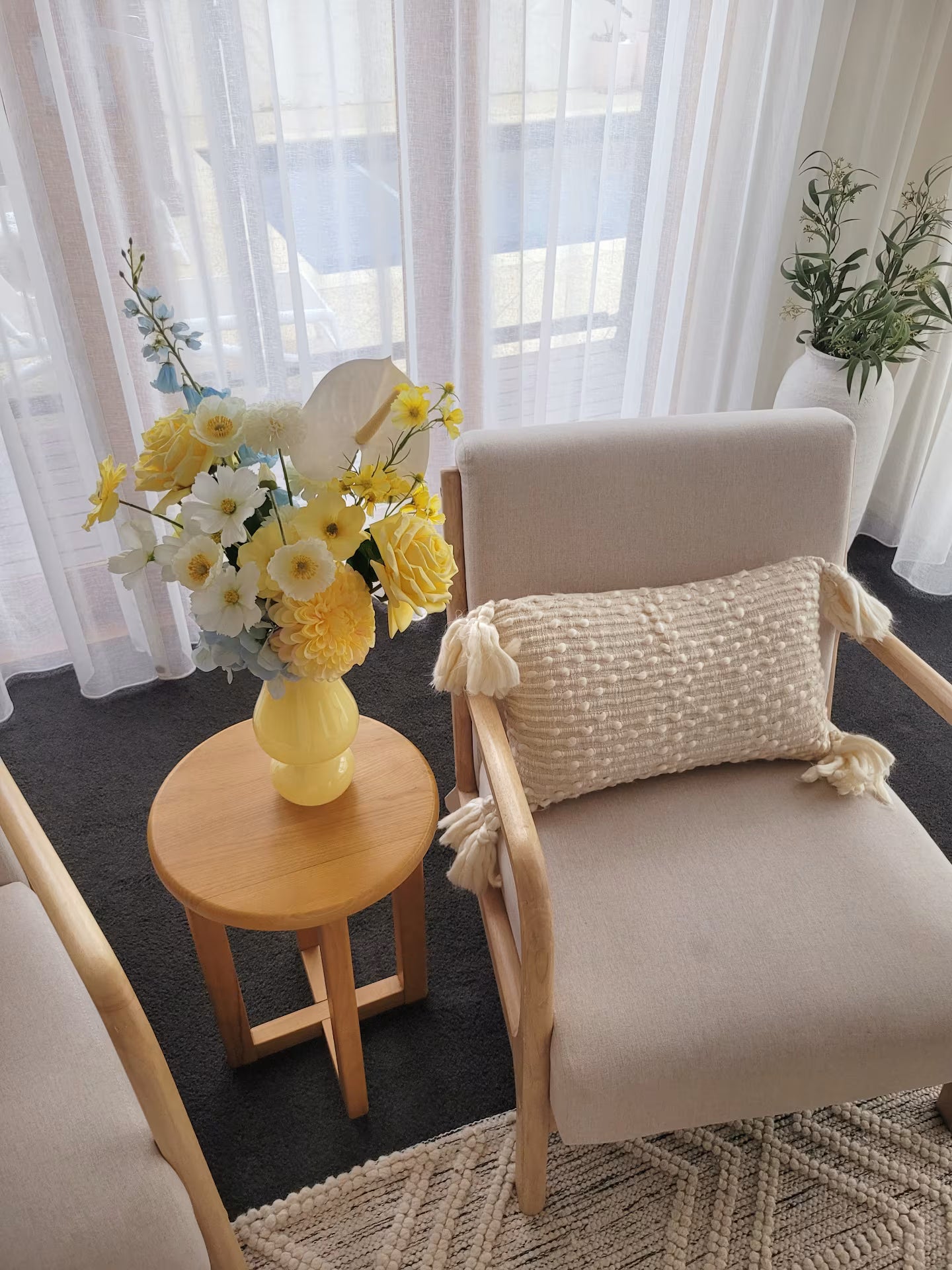Beige armchair with a textured pillow, small round table with a yellow vase and flowers, and sheer curtains in a bright room.