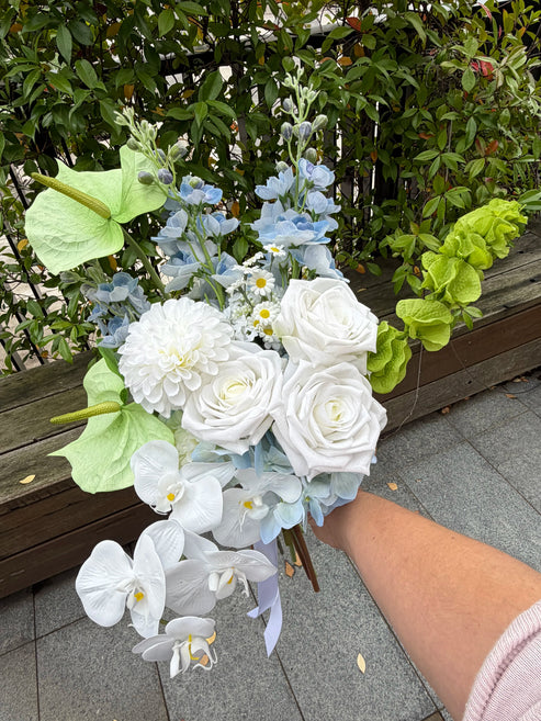 Modern wedding bouquet of white and blue flowers held by a person outdoors.