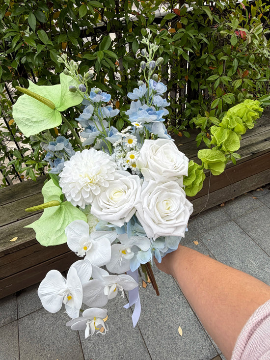 Modern wedding bouquet of white and blue flowers held by a person outdoors.