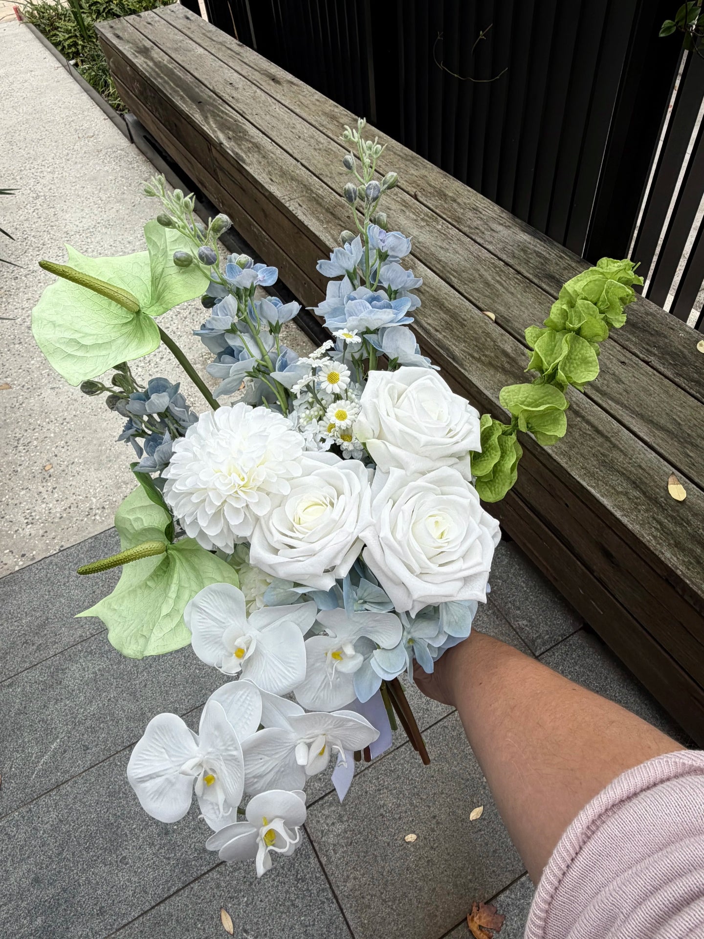Bouquet of white and light blue flowers held by a person on a sidewalk with a wooden bench in the background.