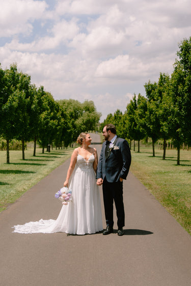 Wedding couple with pastel bouquet standing on a path lined with trees under a cloudy sky.