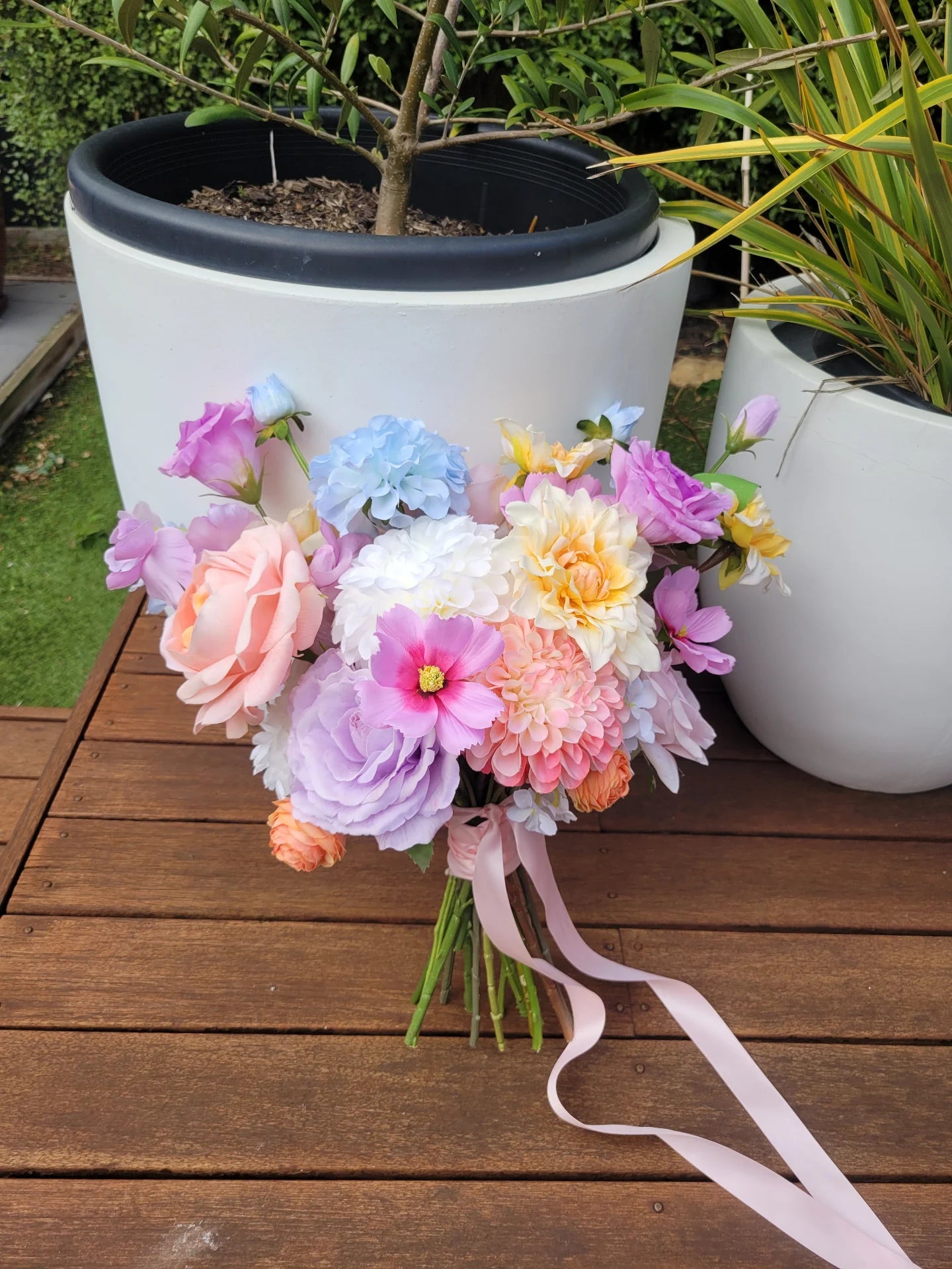 Two wedding bouquets of colourful flowers with a pink ribbon on a wooden surface, with potted plants in the background.