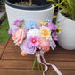Bouquet of colorful flowers with a pink ribbon on a wooden surface, with potted plants in the background.