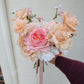 Wedding bouquet of pink and peach flowers held by a person against a white wall.