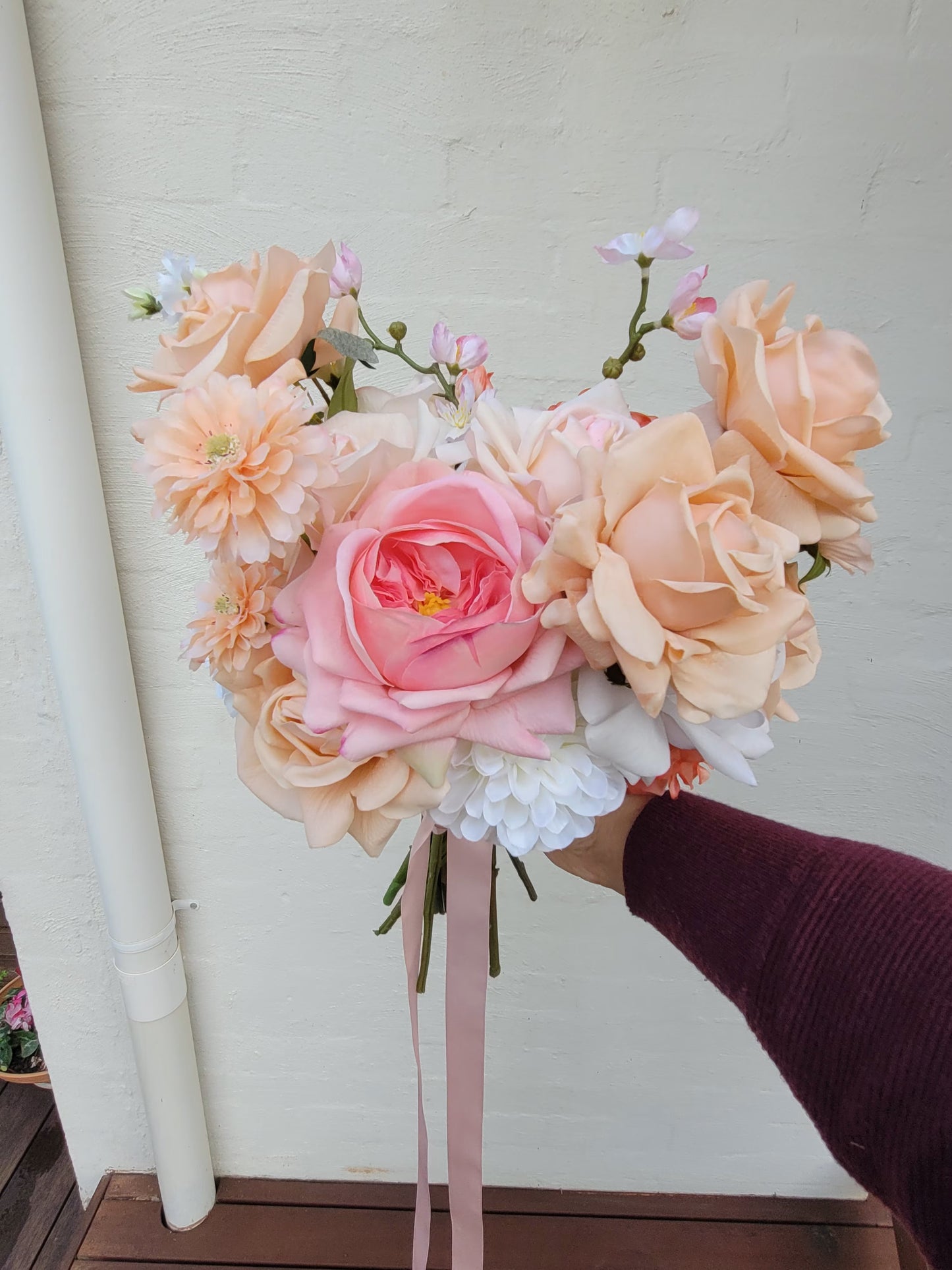 Wedding bouquet of pink and peach flowers held by a person against a white wall.