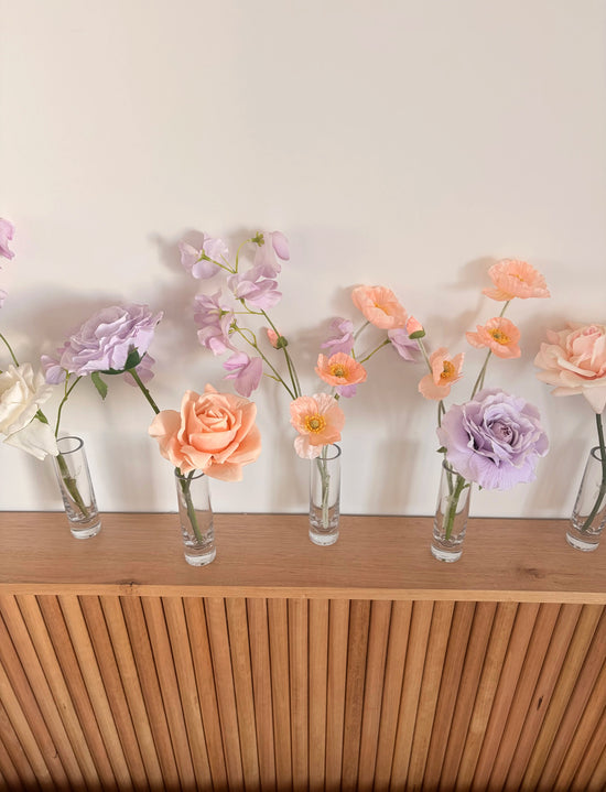 Peach and lilac artificial flower arrangements in small glass bud vases on a wooden surface with a light background