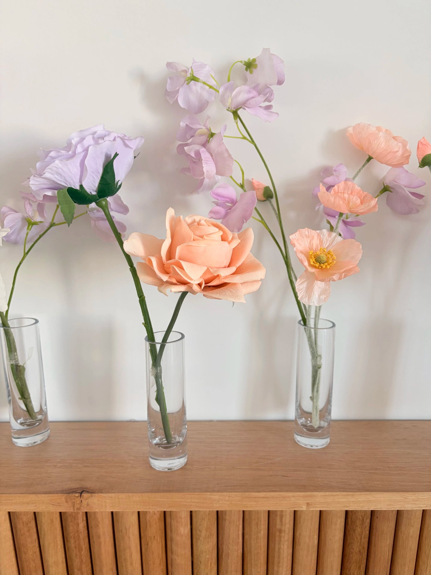 Three glass bud vases with pink and lilac flower arrangements on a wooden surface against a white wall.