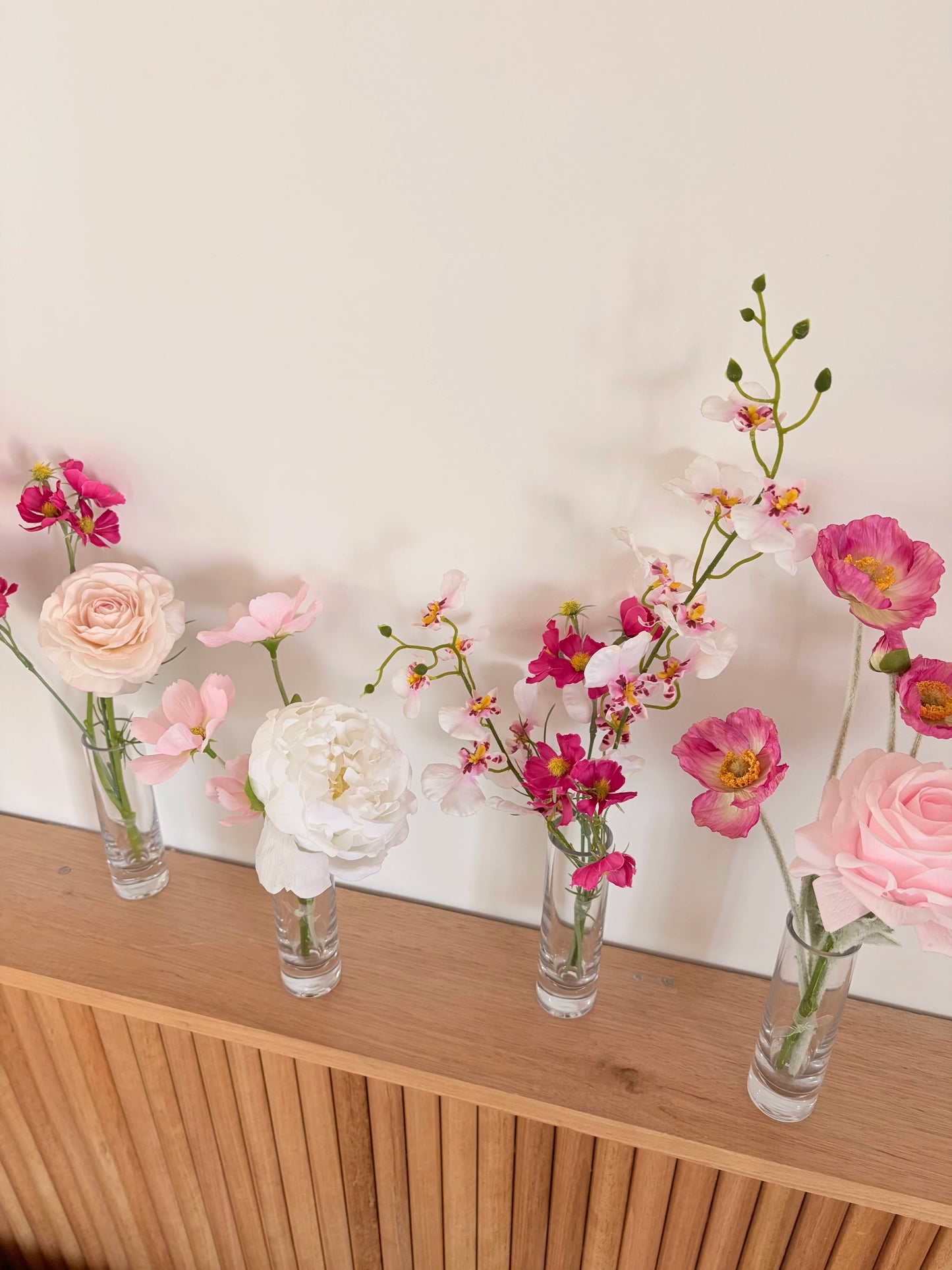 Small glass bud vases with pink and white flowers on a wooden surface against a white wall.
