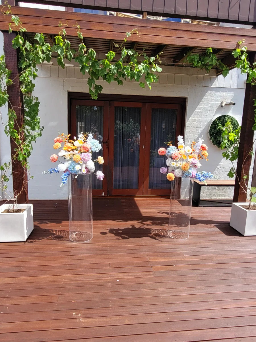 Wedding flowers on clear plinths on a wooden deck with plants and a building in the background.