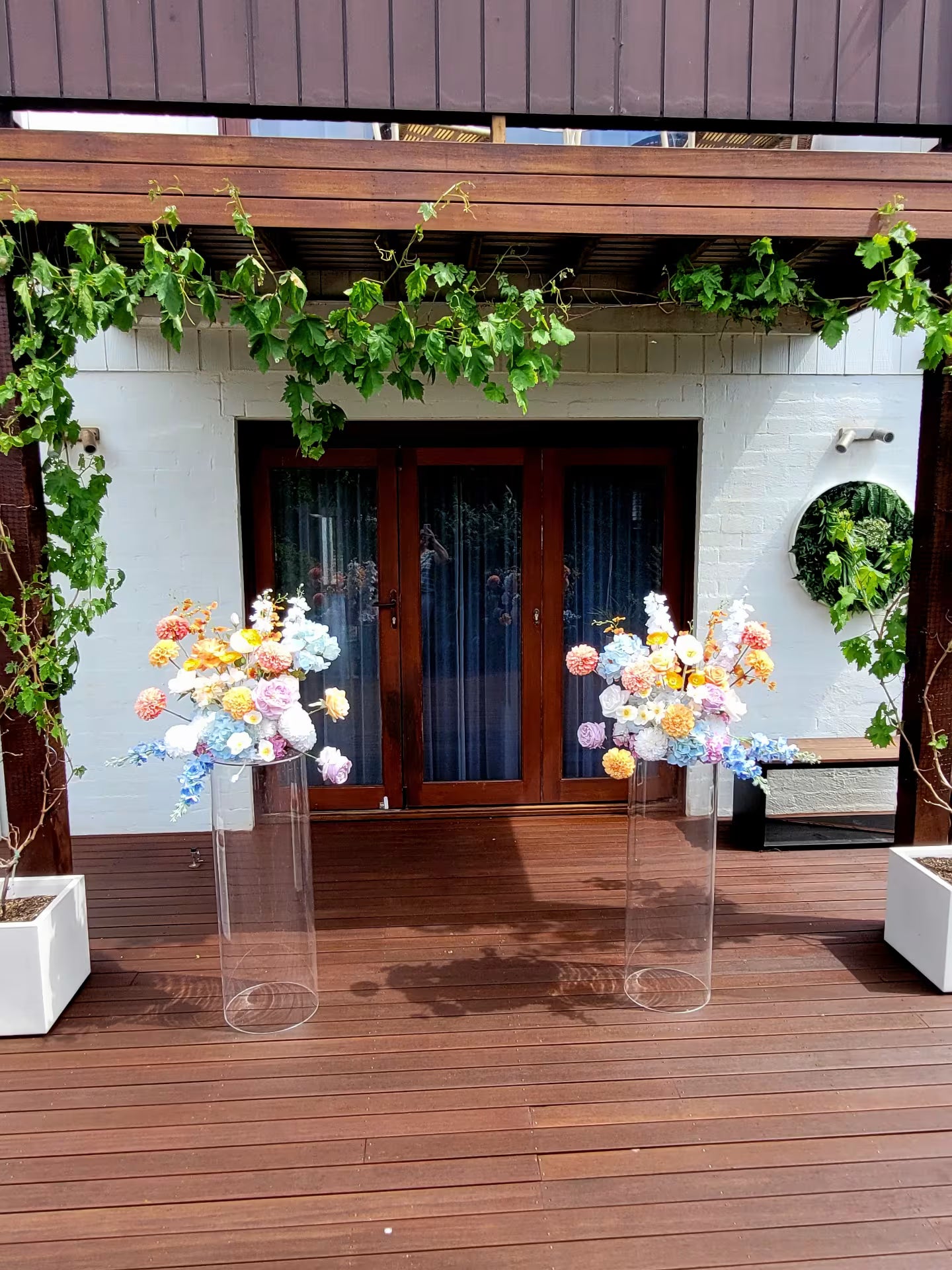 Decorative flowers on clear stands in front of a building with wooden deck and greenery.