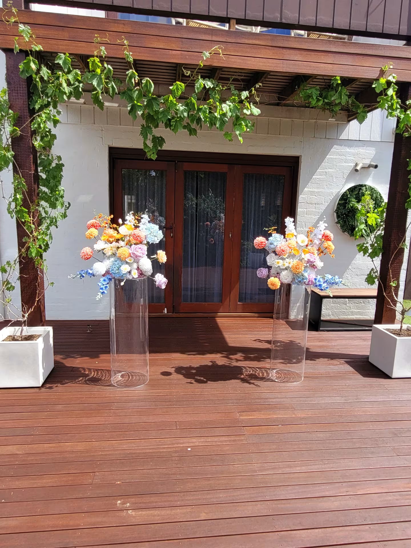 Wedding flowers on clear stands in a backyard setting with wooden deck and plants.