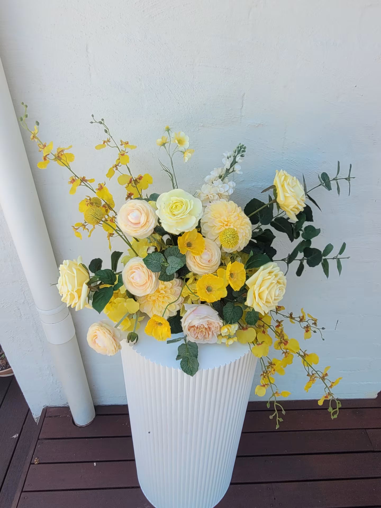 White plinth with yellow and white flowers on a wooden deck against a white wall.