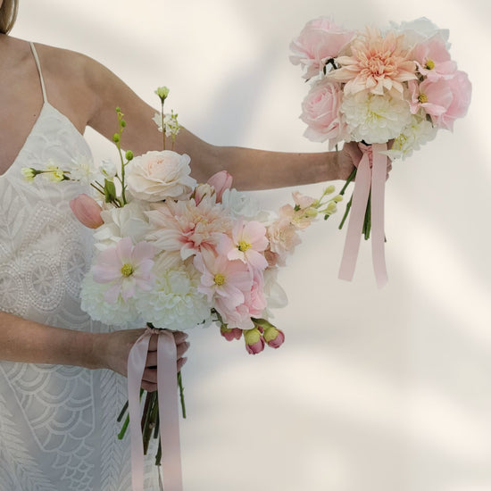 Two wedding bouquets of pink and white flowers held by a person in a white dress against a light background.