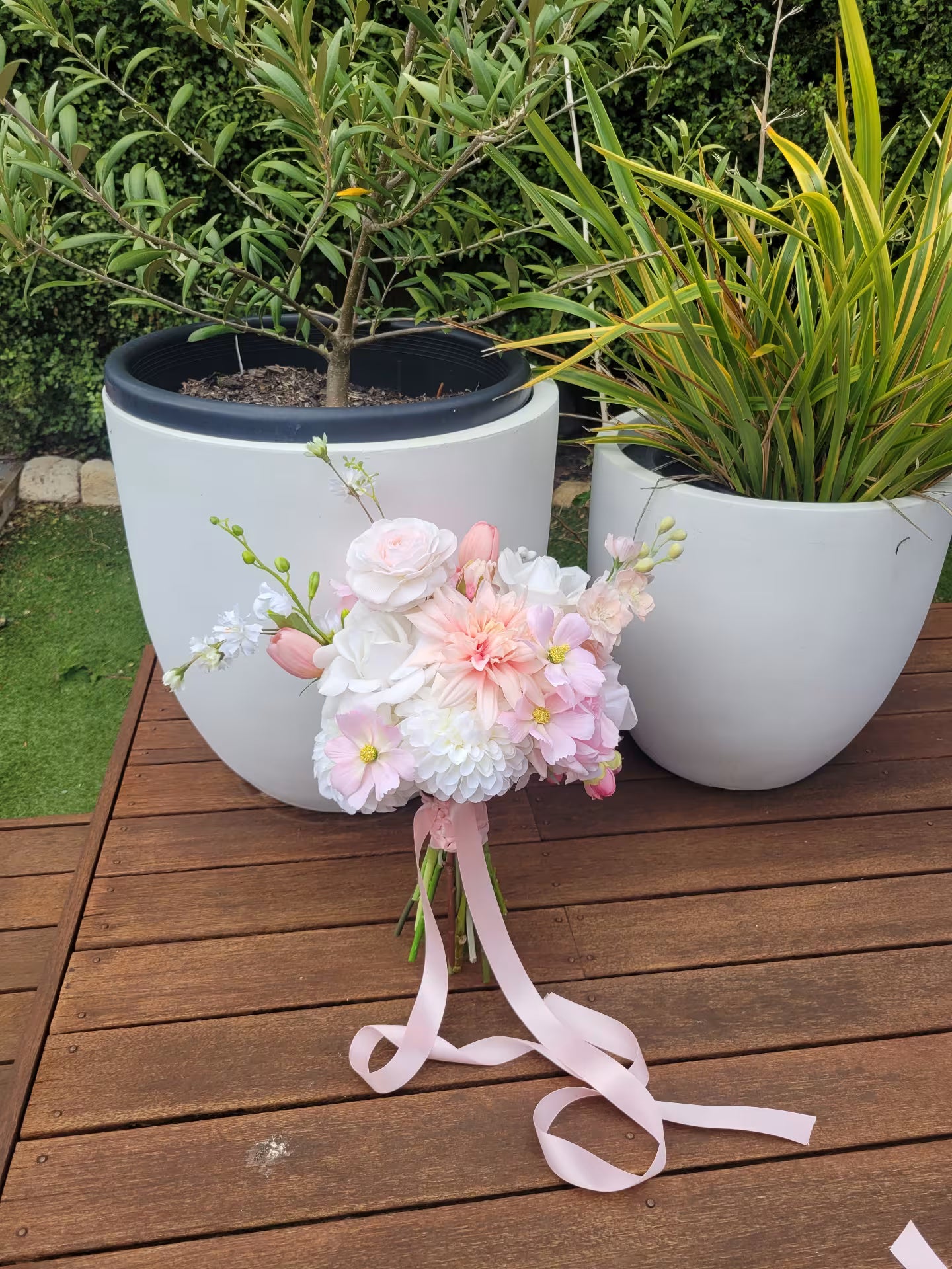 Bridal bouquet of pink and white flowers with ribbons on a wooden table with potted plants in the background.